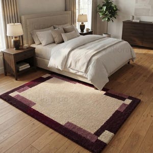 May include: A rectangular area rug featuring a large cream-colored center panel surrounded by a thick border in shades of deep burgundy and muted mauve. The rug has a textured, plush pile surface and is positioned on a light-colored wooden floor.