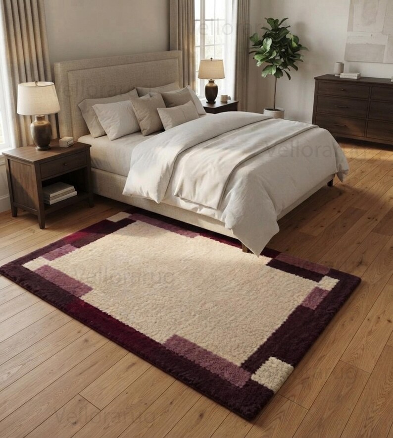 May include: A rectangular area rug featuring a large cream-colored center panel surrounded by a thick border in shades of deep burgundy and muted mauve. The rug has a textured, plush pile surface and is positioned on a light-colored wooden floor.