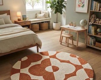 May include: A round area rug featuring a repeating geometric floral pattern in burnt orange and cream. The rug has a textured, tufted pile surface and is positioned on a light-colored wooden floor in a room with a bed, desk, and bookshelf.