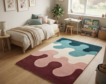 May include: A rectangular area rug featuring a wavy, abstract pattern in layered blocks of deep burgundy, soft pink, cream, and teal blue. The rug is placed on a light-toned wooden floor in a room with a bed, desk, and wall art.