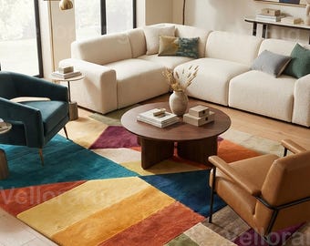 May include: A large rectangular area rug featuring a geometric pattern of color-blocked sections in shades of teal, yellow, orange, red, olive green, and beige. The rug is positioned on a light wood floor beneath a round wooden coffee table.