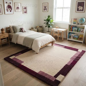 May include: A rectangular area rug featuring a large cream-colored center panel framed by a wide border of deep burgundy and muted mauve geometric blocks. The rug has a textured, plush pile and is positioned on a light-toned wooden floor in a bedroom.