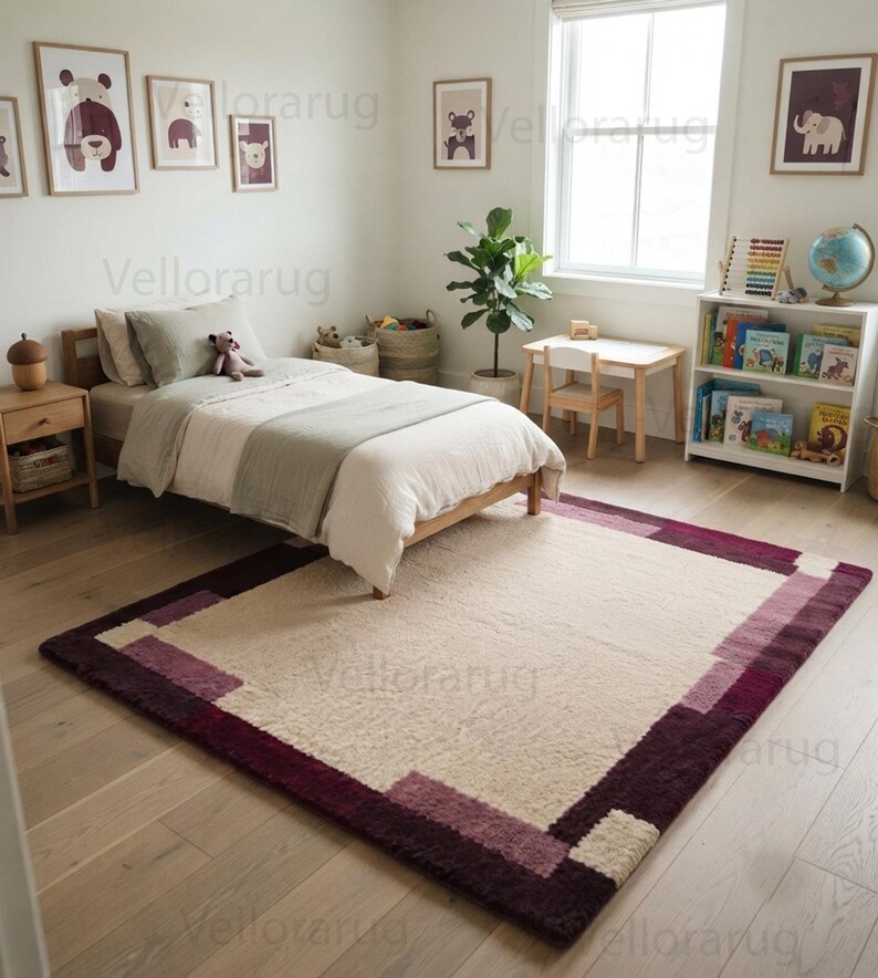 May include: A rectangular area rug featuring a large cream-colored center panel framed by a wide border of deep burgundy and muted mauve geometric blocks. The rug has a textured, plush pile and is positioned on a light-toned wooden floor in a bedroom.