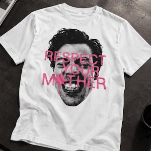 May include: A white short-sleeved t-shirt featuring a black and white photographic print of a person's face with a wide smile. Overlaid on the face is the text "RESPECT YOUR MOTHER" printed in a bold, bright pink font with a star graphic.
