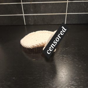 Könnte beinhalten: Ein rundes, cremeweißes gehäkeltes Pad mit strukturierter Oberfläche. Ein schwarzes rechteckiges Etikett mit dem weißen Text "censored" ist diagonal über die Mitte des gehäkelten Objekts gelegt und verdeckt teilweise das runde Textil.