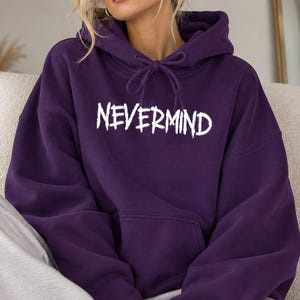 May include: A deep purple hooded sweatshirt featuring a front kangaroo pocket, long sleeves, and a drawstring hood. The word NEVERMIND is printed across the chest in a white, distressed, hand-drawn style font with jagged edges.