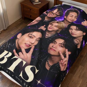 May include: A rectangular blanket featuring a large-scale photographic print of the seven members of the music group BTS. The design includes a deep purple cosmic background with sparkling stars and the large white text BTS at the bottom.