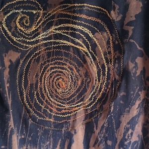 May include: A sleeveless tank top featuring a dark navy blue fabric with a bleached, mottled brown pattern. The front is decorated with a large, spiraling design created with thick, golden-yellow and orange zigzag embroidery stitching.