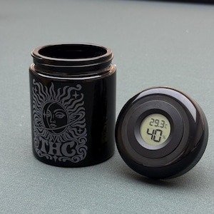 May include: A black glass storage jar with a sun and moon engraving and the letters THC, accompanied by a matching black lid featuring a built-in digital hygrometer display showing 29.3 degrees Celsius and 40 per cent humidity levels.