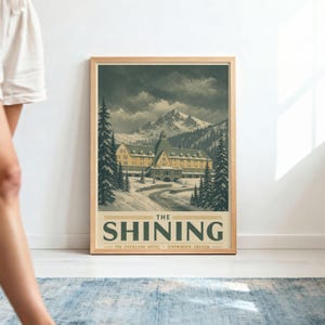 Könnte beinhalten: Ein gerahmtes Poster mit einer verschneiten Berglandschaft, einem großen Hotelgebäude, immergrünen Bäumen und einer kurvenreichen Straße. Der Text lautet THE SHINING, THE OVERLOOK HOTEL, SIDEWINDER, OREGON in dunkelgrüner Schrift auf cremefarbenem Grund.