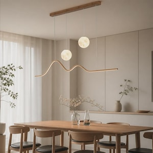 Może przedstawiać: Liniowa lampa wisząca z poziomym drewnianym mocowaniem, dwiema świecącymi kulistymi lampami przypominającymi księżyc oraz cienkim, falistym metalowym prętem w złotym odcieniu, który imituje sylwetkę góry, zawieszona nad drewnianym stołem.