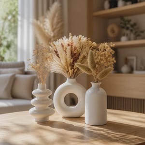 Peut inclure: Trois vases en céramique blanc mat de formes variées contenant des arrangements botaniques séchés. Le vase de gauche a une silhouette ondulée empilée, le vase central est circulaire avec un centre creux, et le vase de droite est une bouteille cylindrique.
