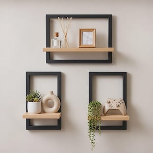 May include: Three wall-mounted shelves featuring black rectangular metal frames and light wood planks. The top shelf holds a glass diffuser, reed sticks, and a wooden frame. The bottom shelves display a succulent, a ceramic vase, and a game controller.