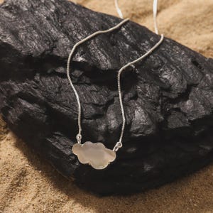 Può includere: Una collana a catena color argento con un ciondolo centrale a forma di nuvola. Il ciondolo ha una superficie liscia, iridescente e bianco sporco con un sottile riflesso. La catena sottile si collega al ciondolo a nuvola in due punti.