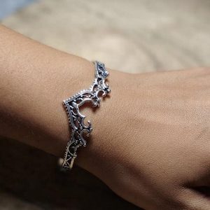 Puede incluir: Una pulsera de brazalete de metal plateado con un intrincado diseño de filigrana calada con un pico central puntiagudo en forma de corona. La pulsera tiene un acabado pulido con áreas empotradas oscurecidas que resaltan los patrones de volutas.
