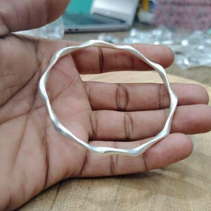 Peut inclure: Un bracelet jonc de couleur argentée présentant un design ondulé et sinueux continu. La surface métallique possède une finition mate et lisse, et l'anneau circulaire conserve une épaisseur constante sur toute sa circonférence.