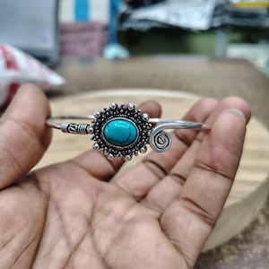 Könnte beinhalten: Ein silberfarbenes Armband mit einem zentralen ovalen Türkisstein, der in einen dekorativen Rahmen im Sonnenstrahlen-Stil gefasst ist. Das Metallband weist auf einer Seite der Steinfassung filigrane Gravuren und ein Spiralmotiv auf.