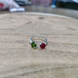 Op de afbeelding: Een open zilveren ring met twee rond geslepen edelstenen in vier-klauwzettingen. De ene edelsteen is levendig groen, terwijl de andere dieprood is. De ringband is glad en gepolijst, waardoor er een duidelijke opening tussen de twee stenen zit.