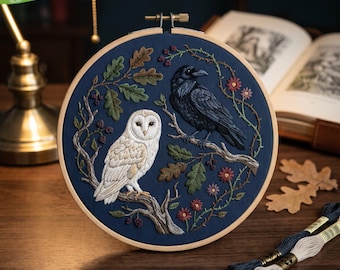 May include: A circular wooden embroidery hoop containing a dark blue fabric panel. The embroidery features a white barn owl and a black raven perched on detailed tree branches, surrounded by green oak leaves, small red berries, and flowers.