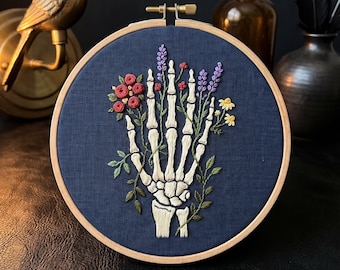 May include: A circular wooden embroidery hoop containing dark blue fabric with a hand-stitched design of a white skeleton hand holding a bouquet of colorful flowers including red roses, purple lavender, and small yellow blossoms with green leaves.