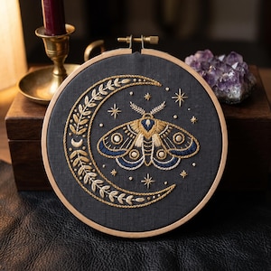 May include: A circular wooden embroidery hoop containing black fabric with hand-stitched gold and navy blue thread. The design features a large crescent moon decorated with leaf patterns, a central moth, and several small star shapes scattered around.