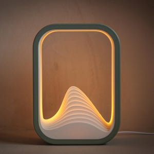 May include: A rectangular table lamp with a sage green frame and a white interior structure featuring layered, wave-like contours. The lamp emits a warm, soft glow from within the center, illuminating the stacked white curves against the frame.