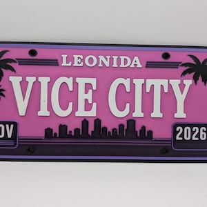 Può includere: Una targa rettangolare con sfondo rosa e bordo viola scuro. Il testo riporta LEONIDA in alto e VICE CITY in grandi lettere bianche. Sagome di palme nere fiancheggiano i lati, insieme a uno skyline cittadino e alla scritta NOV 2026.