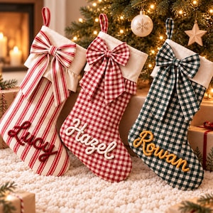 May include: Three Christmas stockings with hanging loops and decorative bows. The first is red and white striped with the name Lucy in red. The second is red and white gingham with the name Hazel in white. The third is green and white gingham with the name Rowan in yellow.