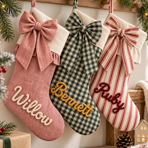 Puede incluir: Tres calcetines navideños con puños y lazos de color crema. El calcetín izquierdo es de color rosa empolvado con el nombre Willow en blanco. El central es de cuadros verdes y blancos con el nombre Bennett en amarillo. El derecho es de rayas rojas y blancas con el nombre Ruby en rojo.