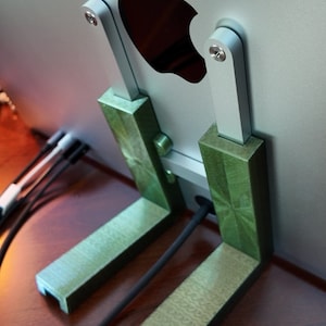 Puede incluir: Un monitor de ordenador plateado con un logotipo de Apple negro está conectado a un soporte verde. El soporte tiene dos soportes verticales conectados por una barra horizontal. El monitor está sentado sobre una superficie de madera.