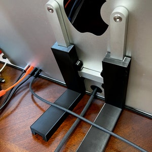 Puede incluir: Un soporte de monitor de computadora impreso en 3D de color negro. El soporte tiene dos soportes verticales y dos soportes horizontales. El soporte está diseñado para elevar el monitor y permitir la gestión de cables.