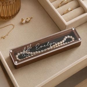May include: A rectangular wooden jewelry box with a clear acrylic lid featuring the white printed name Evelyn, a date of 06.30.2025, a floral sprig, and a heart. Inside the box rests a white pearl necklace against a dark black velvet lining.