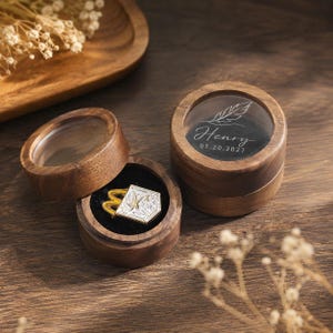 May include: Two round wooden boxes with clear lids. One box is open, revealing a black interior holding a silver-toned pentagonal pin with a gold-colored M logo. The other closed box features a white feather graphic, the name Henry, and 01.20.2027.