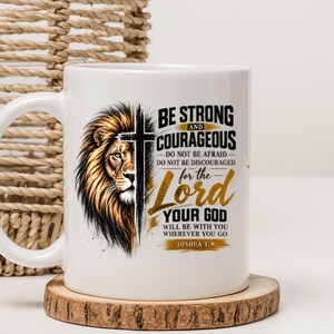 May include: A white ceramic mug featuring a graphic of a lion's face bisected by a black cross. The text reads: BE STRONG AND COURAGEOUS DO NOT BE AFRAID DO NOT BE DISCOURAGED for the Lord YOUR GOD WILL BE WITH YOU WHEREVER YOU GO JOSHUA 1:9.