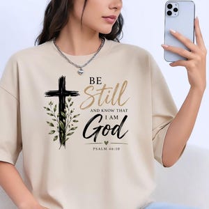 May include: A beige short-sleeved t-shirt featuring a black cross graphic with green leaves and the text "BE Still AND KNOW THAT I AM God PSALM 46:10" in black and gold lettering. The design includes a small heart icon below the word God.