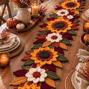 Puede incluir: Un camino de mesa decorativo y alargado con un patrón repetitivo de girasoles y flores blancas más pequeñas tejidas a ganchillo, intercaladas con hojas tejidas en verde oscuro, naranja y rojo intenso, con un diseño floral denso.