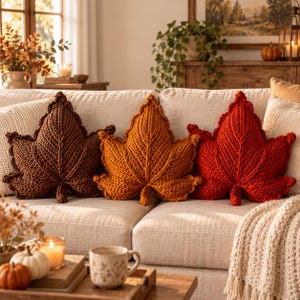 Peut inclure: Trois coussins décoratifs en forme de feuilles d'automne, crochetés en fil marron foncé, orange doré et rouge vif. Chaque coussin présente une nervure centrale détaillée et des bords texturés, disposés côte à côte sur un canapé beige clair.