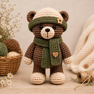 May include: A brown crocheted teddy bear wearing a cream-colored beanie with a green band and a matching green scarf. The bear features cream-colored paws and feet, black eyes, a stitched nose, and a small wooden heart button on the scarf.
