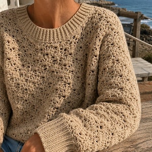 May include: A beige long-sleeved sweater featuring a textured crochet pattern with a repeating shell-like design. The garment has a ribbed crew neckline and ribbed cuffs at the wrists, constructed from a light tan yarn with visible openwork details.