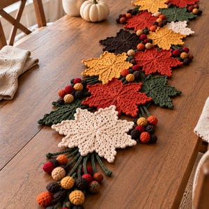 May include: A decorative table runner featuring a series of crocheted maple leaves in shades of cream, yellow, orange, red, dark green, and brown, accented with clusters of small crocheted spheres in matching autumn colors arranged along the length.
