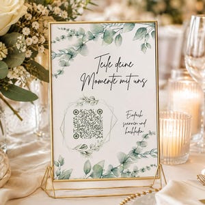 May include: A rectangular sign in a gold metal frame featuring a white background with green eucalyptus leaf illustrations. The text reads Teile deine Momente mit uns and Einfach scannen und hochladen, accompanied by a central QR code graphic.