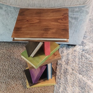 May include: A side table constructed from stacked, painted wooden blocks designed to resemble a vertical pile of books. The table features a dark brown square top, with lower layers painted in shades of green, red, purple, and yellow.