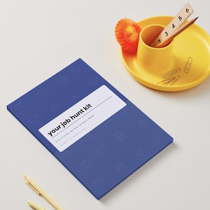 May include: A blue notebook with a white label reading "your job hunt kit" and a pattern of faint icons. Beside it sits a yellow desk organizer containing a wooden ruler marked in inches, a pencil, and paper clips, plus an orange paper flower.