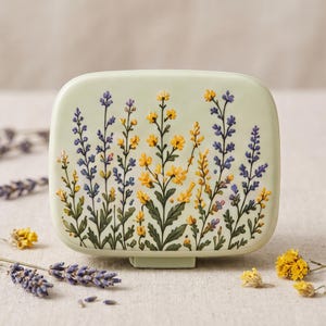 Peut inclure: Un récipient carré vert clair orné d'une illustration en relief de lavande violette et de fleurs sauvages jaunes. Le motif couvre la surface avant avec des tiges, des feuilles et des fleurs détaillées dans des tons de violet, d'or et de vert.