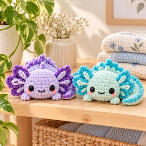 Peut inclure: Deux peluches axolotl crochetées à la main, l'une violet clair et l'autre vert menthe, dotées de grands yeux noirs de sécurité, de joues roses brodées et de structures branchiales détaillées sur les côtés de la tête, posées sur une surface en bois.