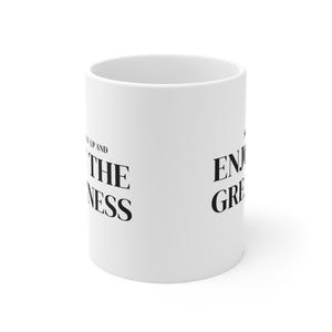 May include: A white ceramic mug featuring black text printed on the side. The visible text reads "UP AND ENJOY THE GREATNESS" in a bold, serif font. The mug has a cylindrical shape with a smooth, matte finish and a standard open top design.