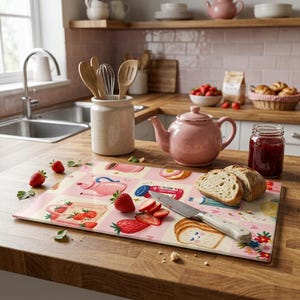 Puede incluir: Una tabla de cortar rectangular de cristal con una cuadrícula de ilustraciones que incluye teteras rosas, bolsas de tela con fresas, tarros de mermelada y rebanadas de pan sonrientes. Un cuchillo de acero con mango blanco descansa sobre ella.