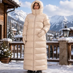 Peut inclure: Un long manteau doudoune matelassé beige doté d'une capuche à col montant, d'une fermeture éclair intégrale sur le devant et de moufles assorties. Le vêtement présente une finition lisse et brillante et descend jusqu'aux chevilles.