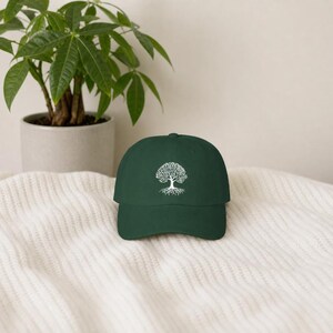 May include: A dark green baseball cap featuring a white embroidered graphic of a tree with visible roots centered on the front panel. The cap has a curved brim and a structured crown, showcasing a clean design with a single color contrast.