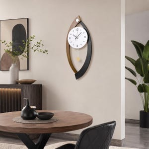 May include: A wall clock featuring a white circular face with black numerals and hands, encased in a vertical, leaf-shaped frame made of two contrasting curved wooden segments in dark brown and black, with a small gold-toned pendulum hanging below.